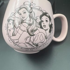 May include: A light pink ceramic mug featuring a black line art illustration of three animated characters enclosed within a circular frame. The mug has a rounded body, a curved handle on the right side, and a smooth, matte-finish exterior surface.