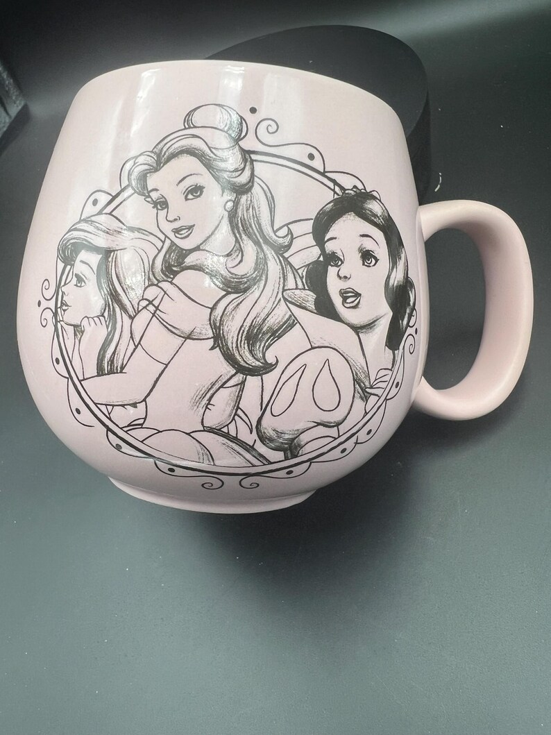 May include: A light pink ceramic mug featuring a black line art illustration of three animated characters enclosed within a circular frame. The mug has a rounded body, a curved handle on the right side, and a smooth, matte-finish exterior surface.