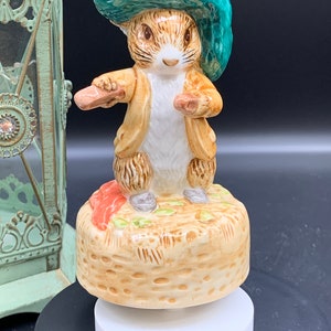 Puede incluir: Una caja de música de porcelana con Peter Rabbit con un sombrero verde y sosteniendo zanahorias. La caja de música está decorada con una base marrón que se asemeja a una cesta.