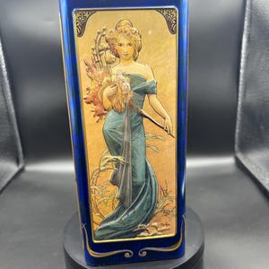 May include: A blue rectangular tin box with a gold trim. The front of the box features a vintage illustration of a woman in a blue dress holding a bouquet of flowers. The illustration is set against a gold background with a decorative border.