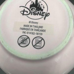 May include: The underside of a light pink ceramic item featuring a black Disney castle logo and text reading Disney, MADE IN THAILAND, and FAC-014182-18159. Two circular icons are present, each with a diagonal line through it, indicating no dishwasher or microwave.