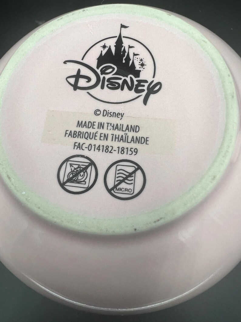 May include: The underside of a light pink ceramic item featuring a black Disney castle logo and text reading Disney, MADE IN THAILAND, and FAC-014182-18159. Two circular icons are present, each with a diagonal line through it, indicating no dishwasher or microwave.