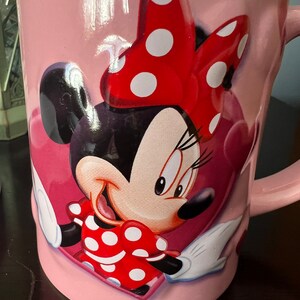 May include: Pink ceramic mug with a cartoon image of Minnie Mouse wearing a red bow with white polka dots. Minnie Mouse is smiling and has a red dress with white polka dots.