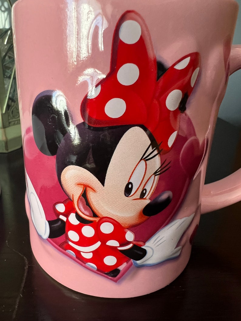 May include: Pink ceramic mug with a cartoon image of Minnie Mouse wearing a red bow with white polka dots. Minnie Mouse is smiling and has a red dress with white polka dots.