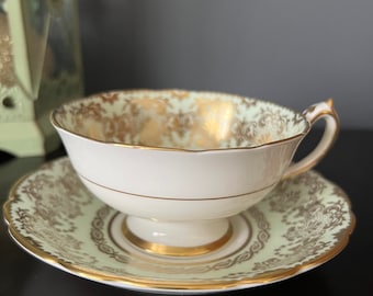 May include: A white teacup and saucer with a green and gold floral pattern. The cup and saucer have a gold rim.