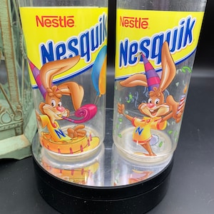 Puede incluir: Dos vasos de plástico transparentes con un logotipo de Nesquik amarillo, azul y rojo y un conejo de dibujos animados con un gorro de fiesta. El conejo sostiene una bocina de fiesta y un pastel con velas. El texto "Nestle" está impreso en los vasos.