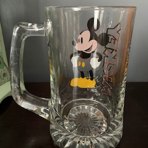 May include: A clear glass beer mug with a handle. The mug has a Mickey Mouse cartoon character printed on the front with the word "Mickey" printed vertically on the right side of the character.