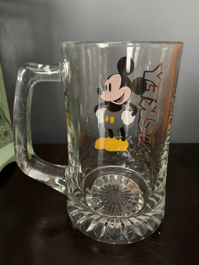 May include: A clear glass beer mug with a handle. The mug has a Mickey Mouse cartoon character printed on the front with the word "Mickey" printed vertically on the right side of the character.