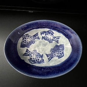May include: A blue and white ceramic oval serving dish with a design of four white fish with black outlines. The dish has a textured blue glaze.