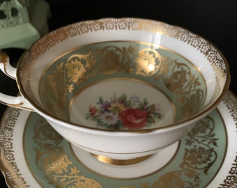Peut inclure: Une tasse à thé et une soucoupe blanches avec un motif floral vert et or. La tasse a une anse et la soucoupe a un bord festonné.
