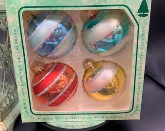 May include: Set of four glass Christmas ornaments with a striped design. The ornaments are hand-decorated with gold and silver glitter and have a white, blue, red, and pink color scheme. The ornaments are packaged in a green box with the text "VERRE DECORE A LA MAIN AVEC COURONNES" and "VIDRIO DECORADO A MANO CON CORONAS".