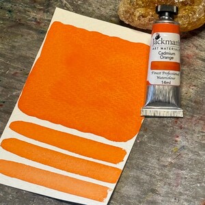 May include: A tube of Jackman's Art Materials Cadmium Orange watercolor paint. The tube is 14 ml and is sitting on a wooden surface with a sample of the paint on a white card.