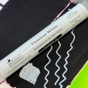 May include: A white Titanium White professional acrylic paint marker with a 3mm round tip, made in England. The marker is shown on a black surface with white paint strokes and a white square. The marker's label includes the brand name and website.