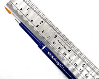 May include: A blue paintbrush with a yellow tip, measuring approximately 7 inches long, with the brand name "michelangelo" printed on the side. The brush is lying on a white surface next to a stainless steel ruler.