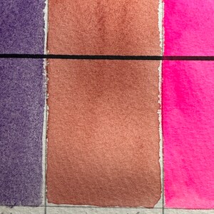 May include: A close-up of a watercolor paint swatch chart. The chart shows three colors: a dark purple, a light brown, and a bright pink. The brown swatch is labeled "Rose Madder".