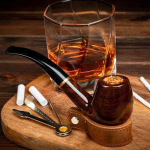 May include: A brown wooden pipe with a leather holder and a bowl filled with tobacco. The pipe is sitting on a wooden cutting board with a glass of amber liquid and a pipe cleaning tool.