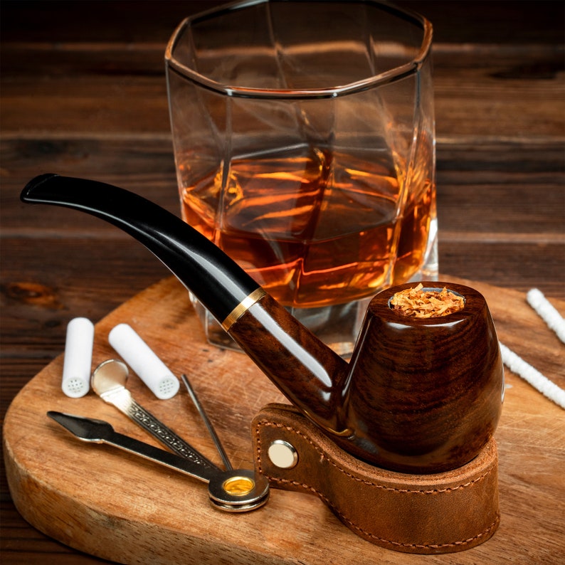 May include: A brown wooden pipe with a leather holder and a bowl filled with tobacco. The pipe is sitting on a wooden cutting board with a glass of amber liquid and a pipe cleaning tool.