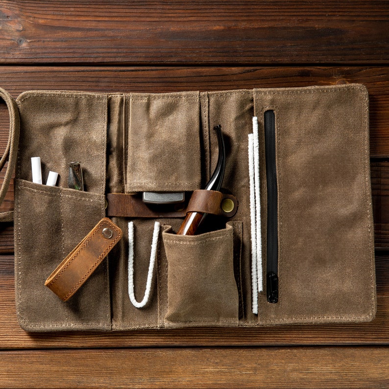 May include: A brown canvas roll-up case with multiple pockets and compartments. The case contains a wooden pipe, a leather pouch, a small metal tool, and several white pipe cleaners.