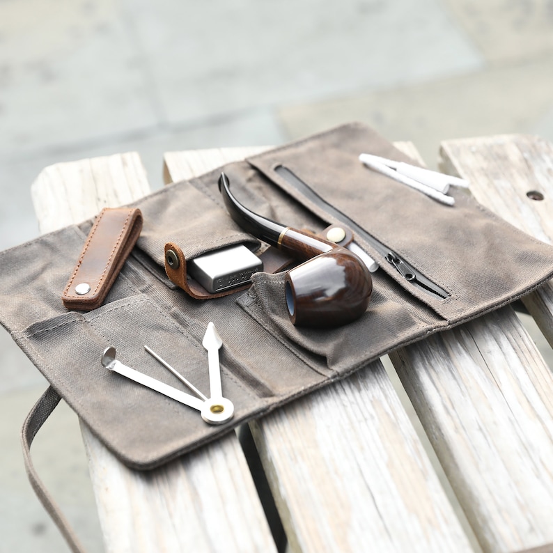 May include: A brown canvas tobacco pouch with a zipper closure. The pouch is open and contains a wooden pipe, a small metal box, and a metal pipe tool.