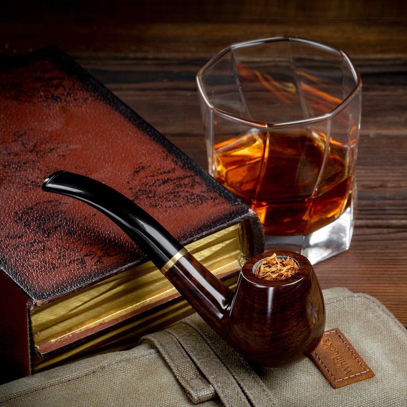 May include: A brown wooden pipe with a bowl filled with tobacco sits on a brown canvas bag. A glass of amber liquid and a brown leather-bound book are in the background.