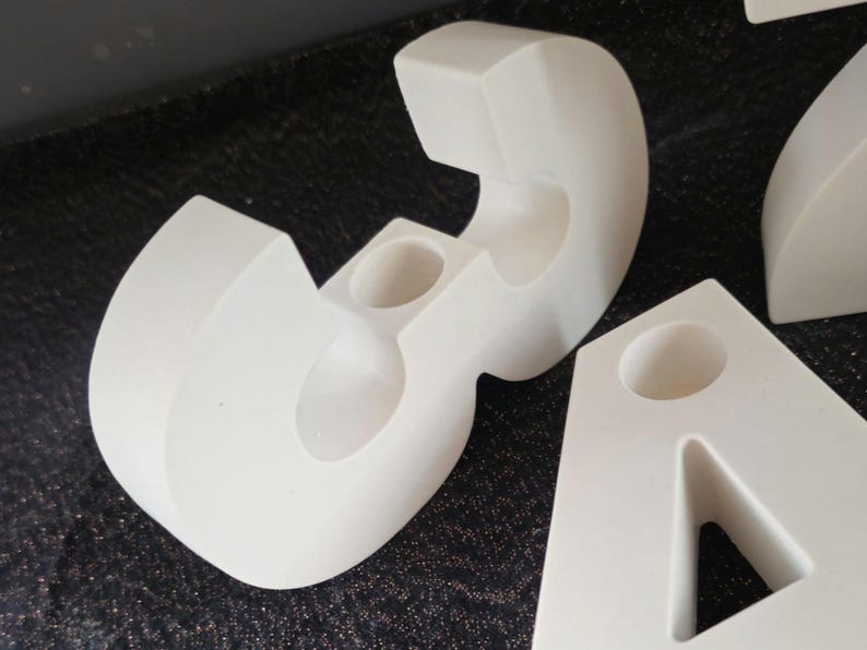 K&ouml;nnte beinhalten: Drei wei&szlig;e Kerzenhalter aus Keramik in Form der Buchstaben C, A und einem stilisierten A. Jeder Halter hat eine kreisf&ouml;rmige &Ouml;ffnung oben f&uuml;r eine Kerze.