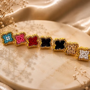 May include: A row of seven square-shaped beaded pins, each featuring a central colour of light blue, pink, red, dark red, black, beige, or white, all framed by a border of metallic gold beads in a uniform, structured square pattern.