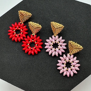 May include: A pair of dangle earrings with a gold-colored metal top and a circle of red and pink beads. The earrings are on a black background.