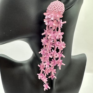 May include: Pink floral dangle earrings. The earrings feature a cluster of small, round beads at the top, transitioning into cascading strands of tiny flowers and beads. The earrings are a soft, pastel pink color.
