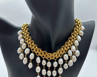 May include: A gold-toned necklace with a chain design and dangling white pearls. The necklace is displayed on a black mannequin, showcasing its layered design and the contrast between the gold and the pearls.