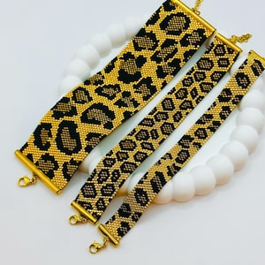 Può includere: Quattro braccialetti di perline con un motivo leopardato nero e oro. I braccialetti sono esposti su una superficie bianca e ondulata. Ogni braccialetto ha estremità e chiusure in metallo color oro.