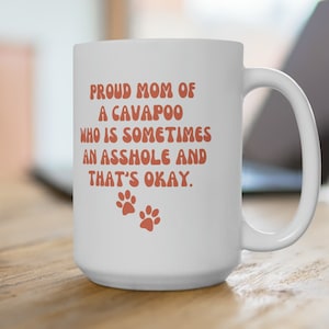 Op de afbeelding: Witte keramische mok met de tekst "PROUD MOM OF A CAVAPOO WHO IS SOMETIMES AN ASSHOLE AND THAT'S OKAY." in oranje letters. Twee oranje pootafdrukken staan eronder. De mok staat op een houten oppervlak.