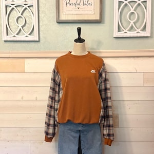 May include: A long-sleeve crewneck sweatshirt featuring a solid burnt orange front panel with a small white Nike logo on the chest and plaid patterned sleeves in shades of blue, white, and orange, paired with light blue denim jeans.