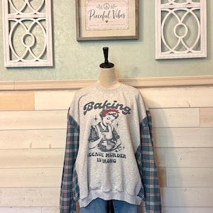 May include: A light gray crewneck sweatshirt featuring a graphic of a person baking with the text "Baking because murder is wrong" in black lettering. The sleeves are made of a blue, green, and red plaid fabric, contrasting with the gray body.