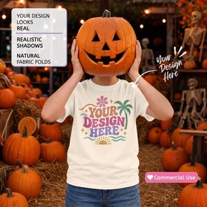 May include: A cream-colored t-shirt featuring a graphic print with a pink flower, a green palm tree, wavy lines, and a yellow sun. The text reads YOUR DESIGN HERE. The shirt is worn by a person holding a carved orange jack-o'-lantern pumpkin.