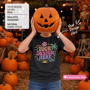 May include: A black t-shirt featuring a graphic with the text "YOUR DESIGN HERE" in pink and purple, surrounded by a flower, a palm tree, waves, and a sun. The shirt is displayed in a setting filled with orange pumpkins and skeletons.