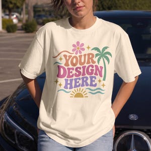 May include: A cream-colored short-sleeve t-shirt featuring a graphic print with the text "YOUR DESIGN HERE" in a wavy font. The design includes a pink flower, a green palm tree, a yellow sun, and small stars in a retro-inspired color palette.