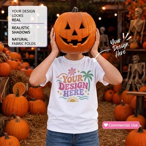 May include: A white t-shirt featuring a colorful graphic with the text "YOUR DESIGN HERE" surrounded by a flower, palm tree, sun, and wavy lines. The shirt is held up by a person hiding their face behind a large, carved orange jack-o'-lantern.