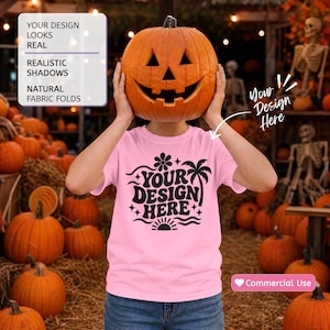 May include: A light pink short-sleeve t-shirt featuring a black graphic print with the text YOUR DESIGN HERE, a palm tree, a flower, and a sun. The shirt is held up by a person wearing a carved orange jack-o'-lantern over their head.