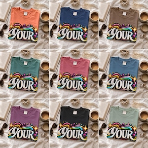 Può includere: Una collezione di nove magliette in vari colori tra cui arancione, blu, marrone, verde, rosa e nero. Ogni maglietta presenta una stampa grafica sul fondo con la parola YOUR in grandi lettere bianche circondata da vortici e stelle colorate.