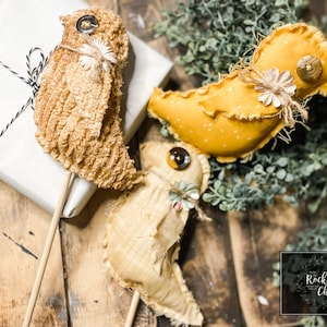 以下が含まれることがあります： 棒につけられた手作り布製の鳥が3羽。鳥は茶色、黄色、白の色合いで、素朴でヴィンテージな雰囲気です。鳥は木製の表面の上に置かれています。