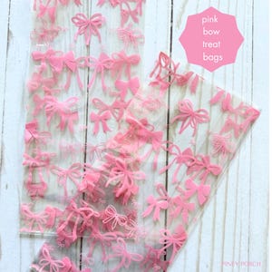 May include: Clear plastic treat bags decorated with pink bows. The bags are filled with multiple pink bow designs. The text "pink bow treat bags" is in a pink hexagon shape. The background is a light wood.