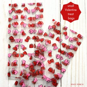 May include: Clear plastic treat bags decorated with red and pink hearts. The bags are filled with heart-shaped confetti. The text "small Valentine treat bags" is in a red octagon.