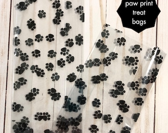 May include: Clear plastic treat bags with a repeating pattern of black paw prints. The bags are rectangular and partially transparent, with the words "paw print treat bags" in a black hexagon.
