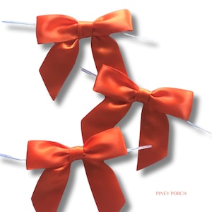 May include: Three orange satin ribbon bows, each featuring a central knot and two downward-pointing tails. Each bow is attached to a thin, flexible wire stem extending from the sides. The text PINEY PORCH is printed in small orange letters.