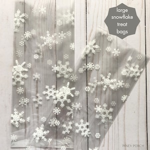 May include: Clear plastic treat bags featuring a white snowflake pattern of varying sizes. A gray hexagonal graphic in the upper right corner contains the text "large snowflake treat bags" and the bottom right corner displays the text "PINEY PORCH".