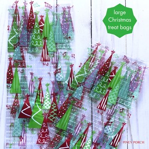May include: A pile of clear cellophane treat bags with a Christmas tree pattern. The bags are printed with red, green, and purple trees with white accents. The text "large Christmas treat bags" is written on a green circle in the upper right corner of the image.