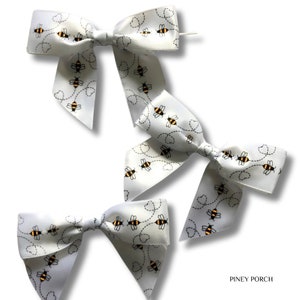 May include: Three white satin bows with a bee and dotted line pattern. The bows are tied with a white ribbon and are perfect for adding a touch of whimsy to any gift or package.