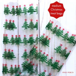 May include: Clear plastic treat bags with a repeating pattern of green Christmas trees with red star and red ball ornaments. The text "medium Christmas treat bags" is printed in red on a white background. The text "Piney Porch" is printed in black at the bottom right corner of the image.