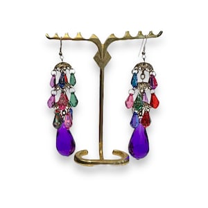 May include: A pair of dangle earrings with a cascade of colorful teardrop-shaped crystals. The earrings are suspended from a gold-toned metal base and feature a large, teardrop-shaped purple crystal at the bottom.