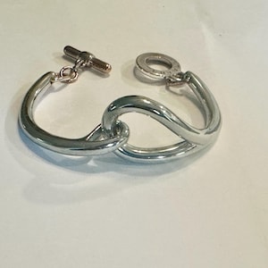 May include: A silver-toned bracelet with a toggle clasp. The bracelet features a looped design, with two curved sections that intertwine. The toggle clasp is a small, cylindrical bar that fits into a circular loop to secure the bracelet.